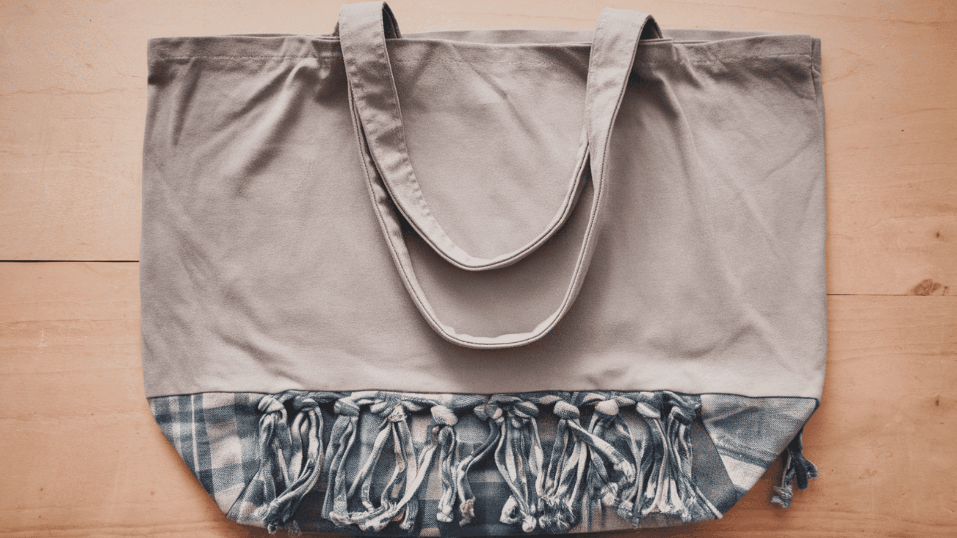 a beige canvas tote bag with denim fringe trim laid flat on a light wooden surface