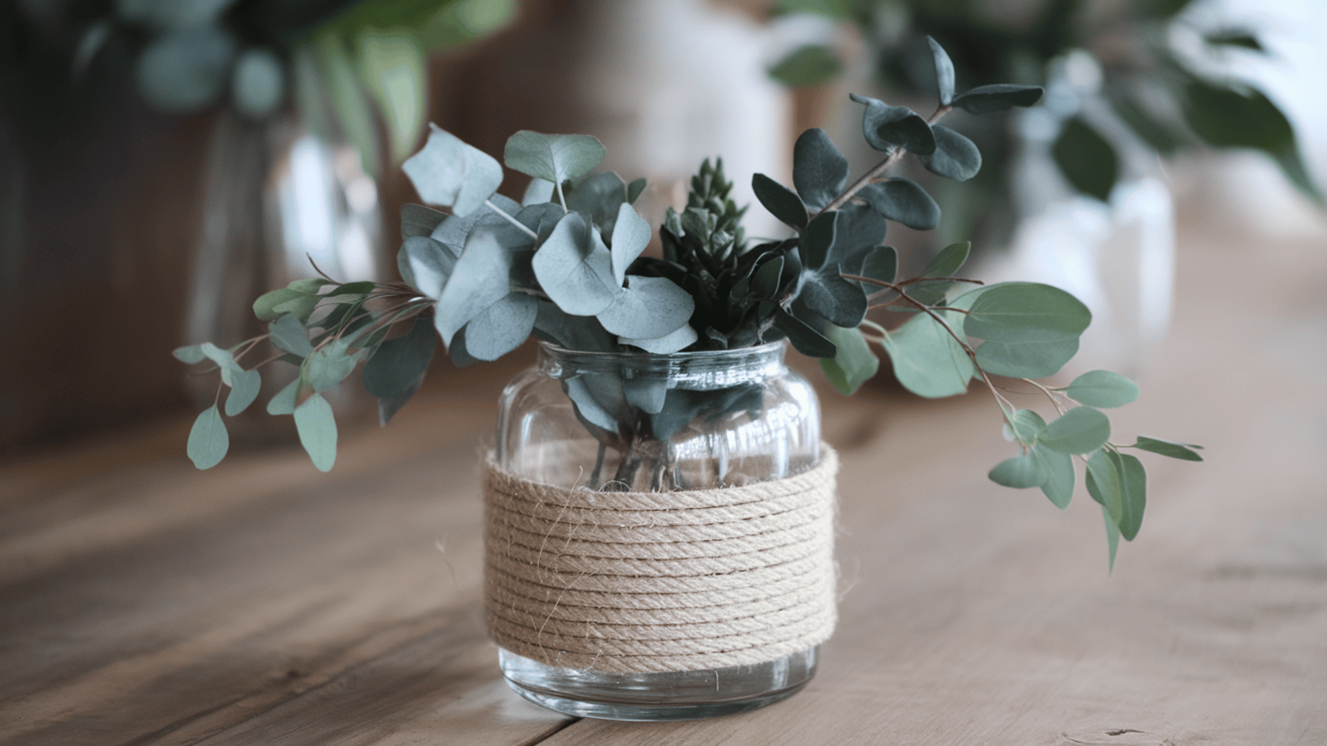 a glass jar wrapped with coiled jute rope holding eucalyptus and succulent stems on a wooden table
