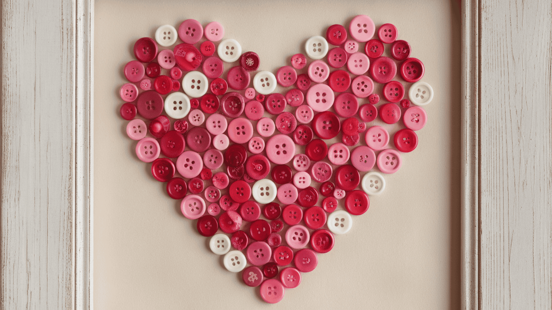 a heart shape made from red, pink and white buttons arranged on canvas inside a white wooden frame