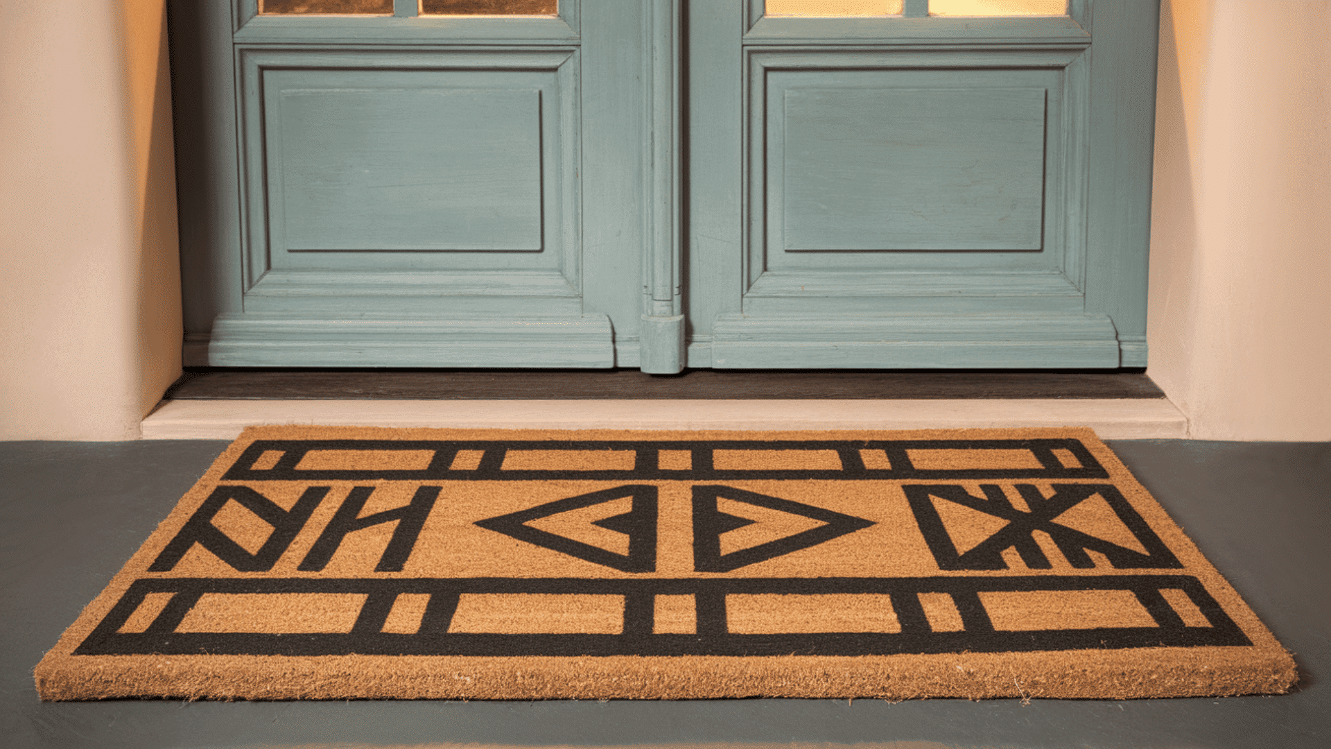 a natural coir doormat with bold black geometric tribal pattern placed in front of a teal double door