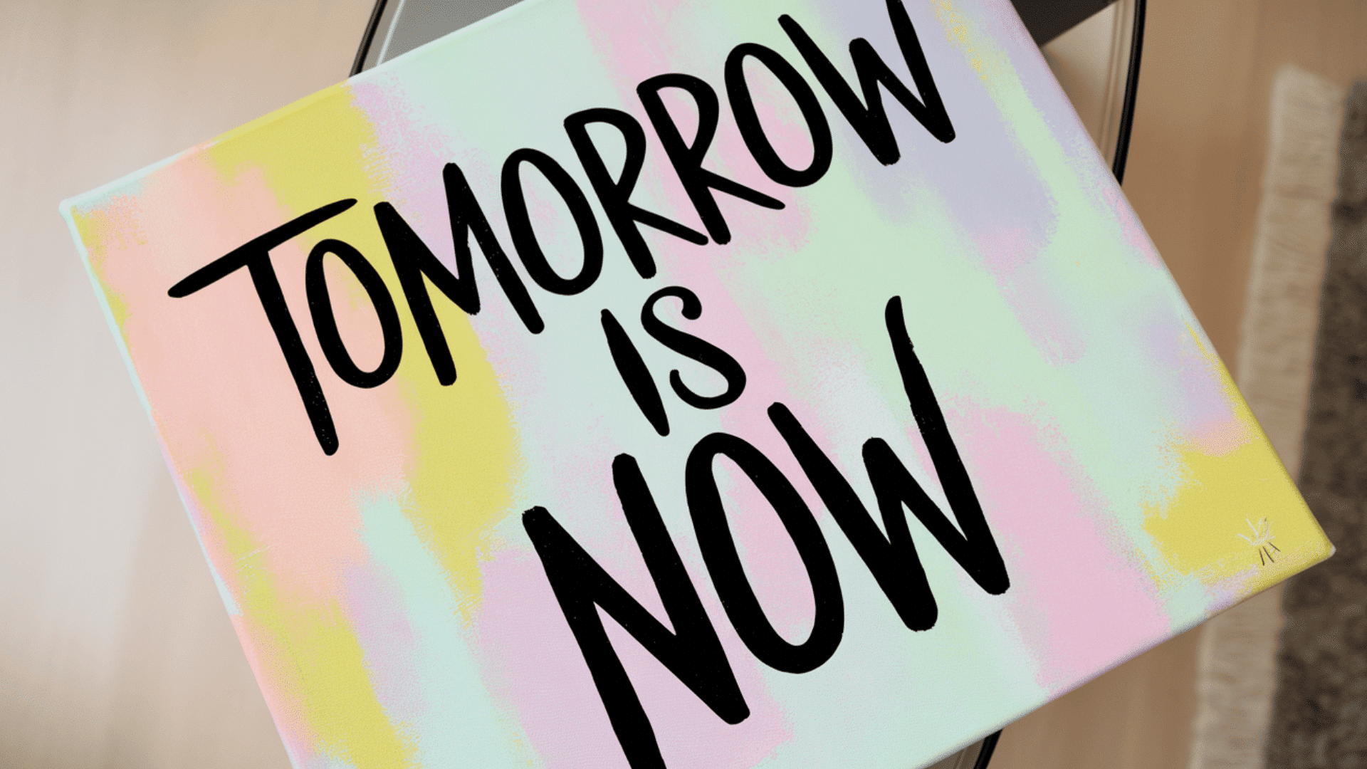 a pastel watercolor canvas with black hand lettered quote reading tomorrow is now leaning on a stand