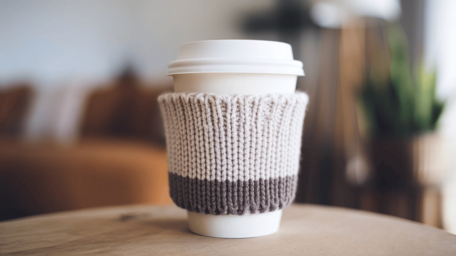 a white takeaway coffee cup with a two tone beige and brown knitted cozy sleeve on a wooden table