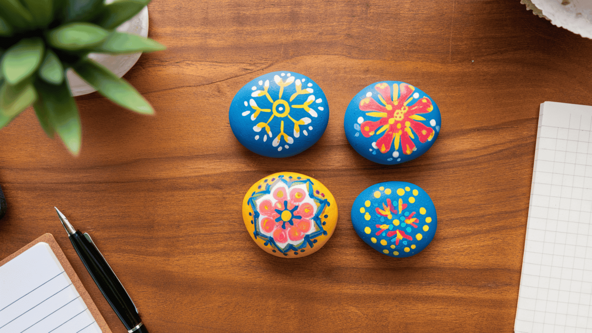 four colorful hand-painted rocks with floral mandala designs placed on a wooden desk with a notepad