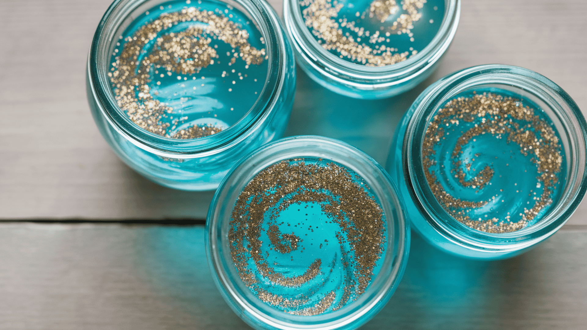 four glass jars filled with teal blue wax and gold glitter swirls viewed from above on a wood surface