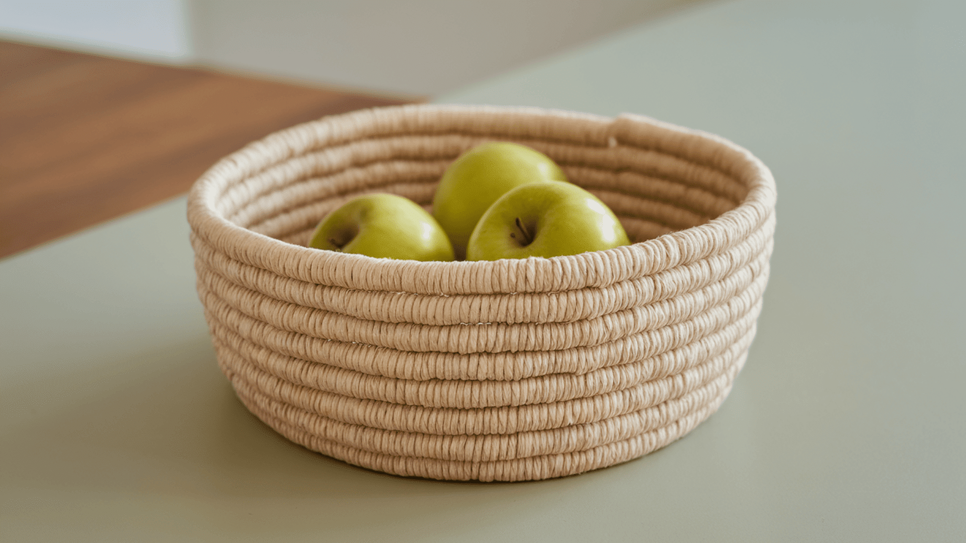 three green apples sitting inside a handmade natural rope coil bowl placed on a sage green surface