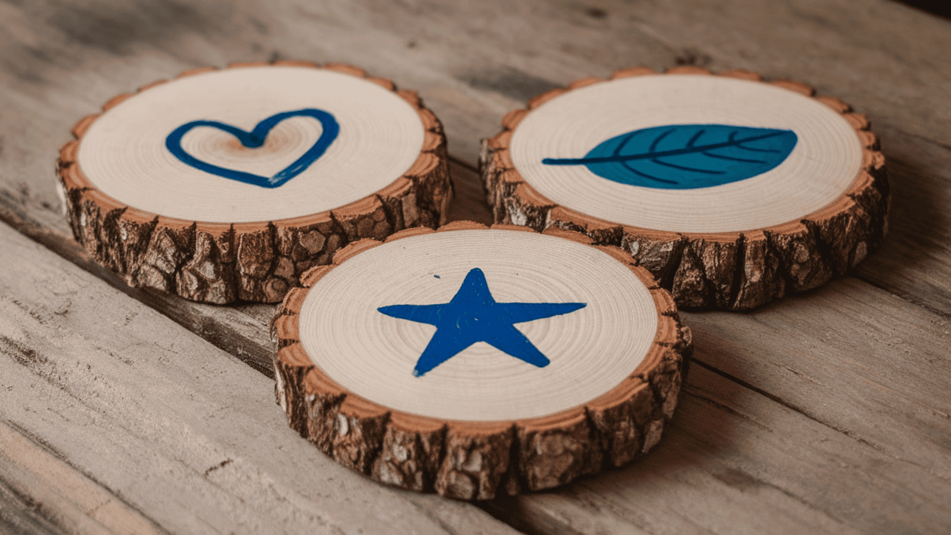 three round wood slice coasters with blue painted heart star and leaf designs on a weathered wood table