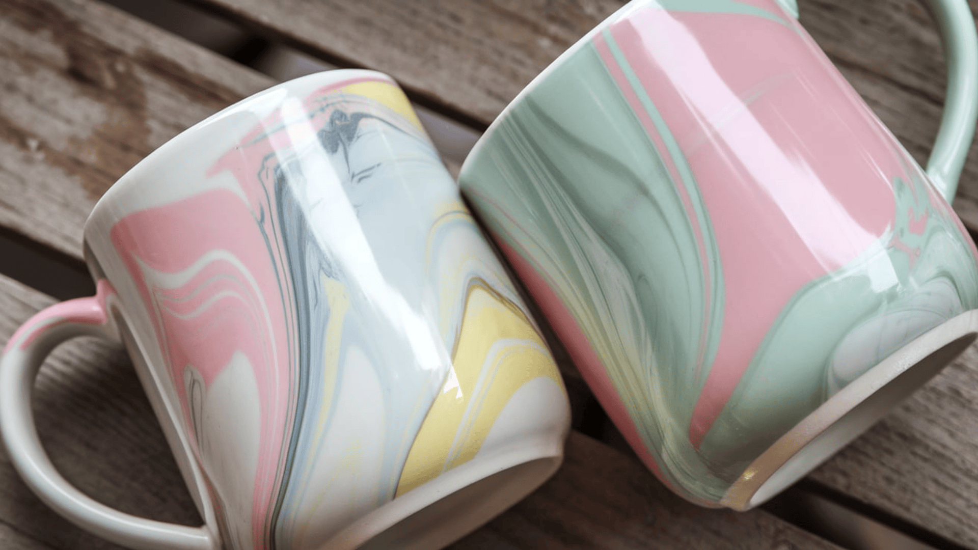 two ceramic mugs with pastel pink yellow and green marble swirl patterns resting on a wooden surface