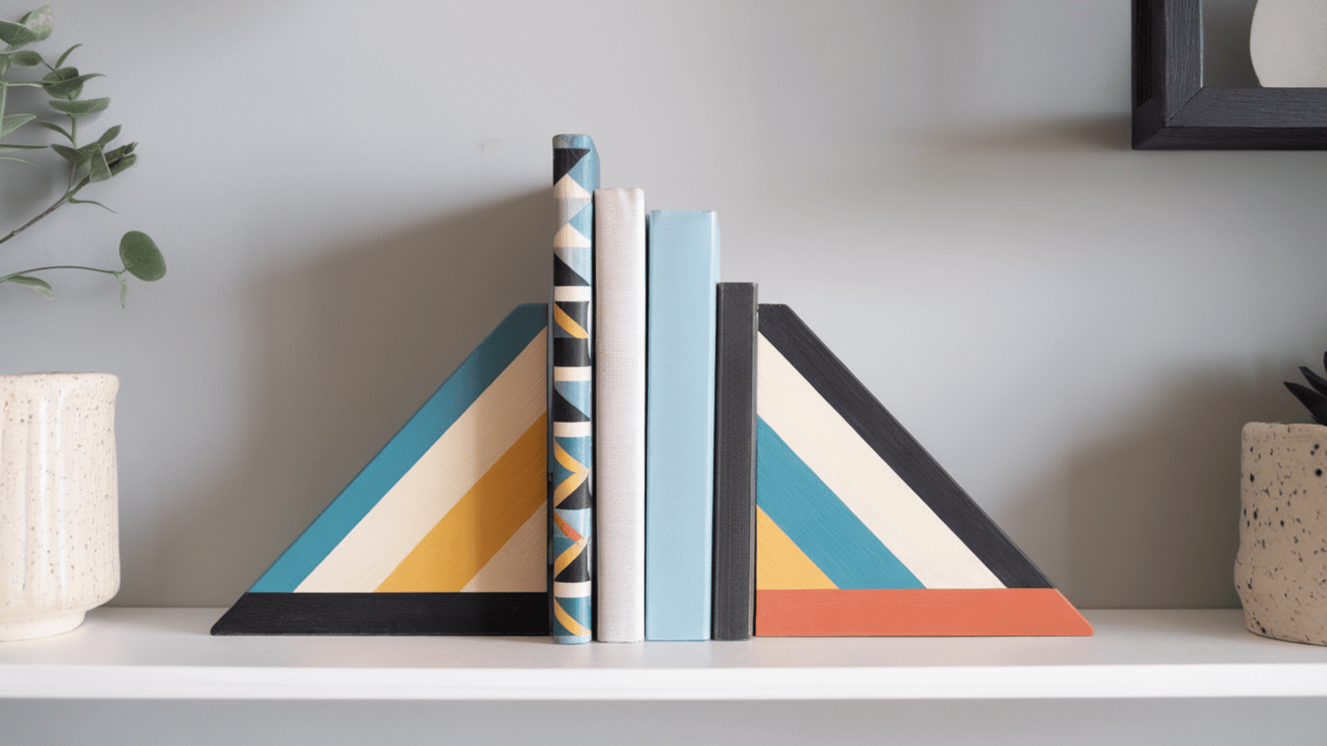 two triangle shaped wooden bookends painted with bold color block stripes holding books on a white shelf